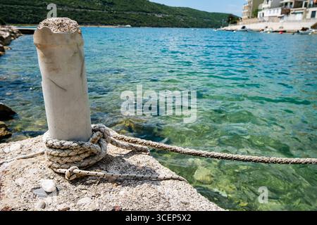 Close-up of a thick rope securely tied around a weathered concrete mooring bollard on a pier, with clear blue sea water in the background. Stock Photo