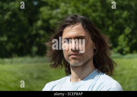 Portrait of handsome caucasian brunette man with long curly hair and dissatisfied emotion and feeling unwell at park and green nature setting. Summer Stock Photo