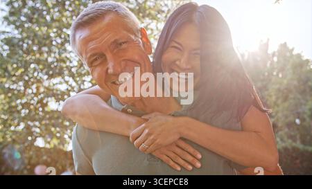 Outdoor Portrait Of Mature Couple With Woman Hugging Man From Behind With Lens Flare Stock Photo