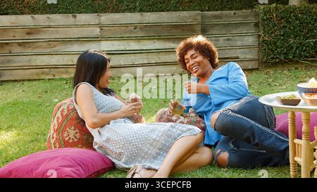 Two Smiling Mature Female Friends Enjoying Drinks And Snacks In Backyard Garden Together Stock Photo