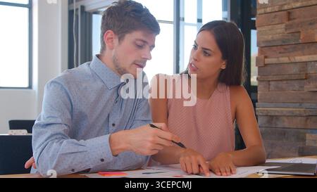Businessman And Businesswoman Sitting At Desk In Open Plan Office Discussing Documents Stock Photo