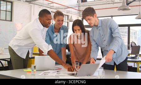 Young Business Team Meeting Standing Around Table With Laptops And Documents Stock Photo