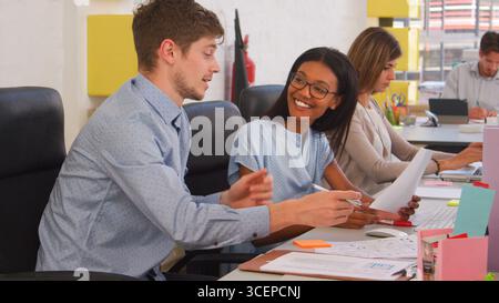 Business Team Sitting At Desks In Open Plan Office Working At Computers And Discussing Documents Stock Photo