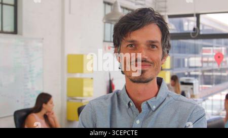 Head And Shoulders Portrait Of Smiling Businessman In Busy Office Stock Photo