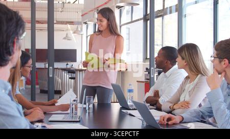 Young Businesswoman Making Presentation To Colleagues Using Laptops Sitting Around Boardroom Table Stock Photo