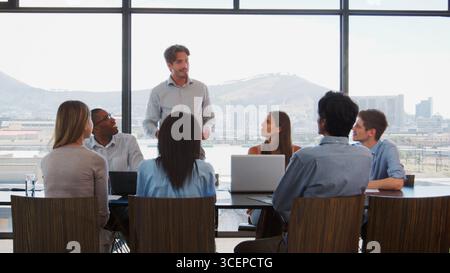 Businessman Making Presentation To Colleagues Using Laptops Sitting Around Boardroom Table Stock Photo