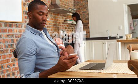 Father Holding Baby Son Whilst Using Laptop As Mother Prepares Meal At Home Stock Photo