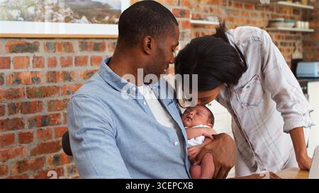 Father Holding Baby Son As Mother Kisses His Head At Home In Kitchen Stock Photo