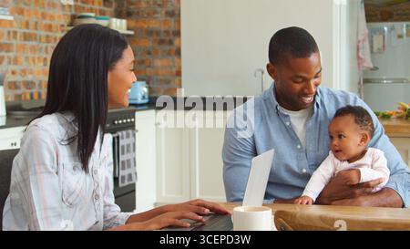 Mother Working On Laptop Whilst Father Sits At Table Cuddling Baby Daughter Stock Photo