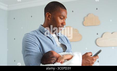 Father Holding Newborn Baby Son Whilst Using Mobile Phone In Nursery At Home Stock Photo