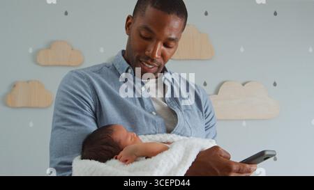 Father Holding Newborn Baby Son Whilst Using Mobile Phone In Nursery At Home Stock Photo