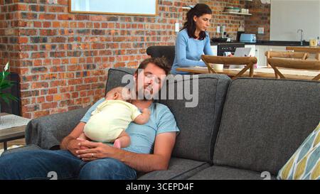 Mother Working On Laptop Whilst Tired Father Sleeps On Sofa Cuddling Baby Son Stock Photo