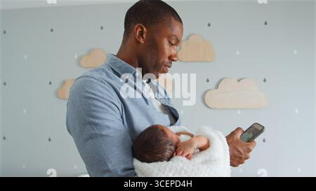Father Holding Newborn Baby Son Whilst Using Mobile Phone In Nursery At Home Stock Photo