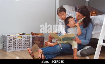 Mother Sitting On Floor Reading Story To Children In Their Bedroom At Home Stock Photo