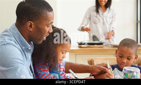 Father And Children Drawing At Table As Mother Prepares Meal Stock Photo