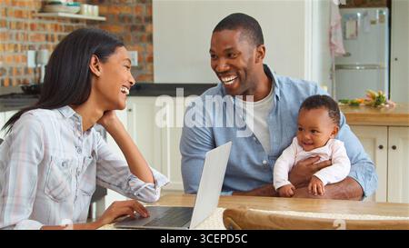 Mother Working On Laptop Whilst Father Sits At Table Cuddling Baby Daughter Stock Photo