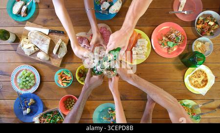 Overhead Close Up Shot Of Friends Eating Meal Around Table Doing Cheers With Beer And Wine Stock Photo