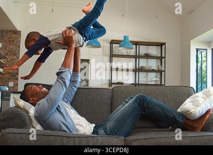 Father Lying On Sofa At Home Having Fun With Son Lifting Him Into The Air Stock Photo