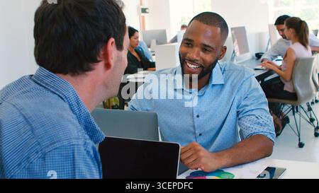 Two Businessmen Having Informal Meeting Working At Desks On Laptops In Modern Open Plan Office Stock Photo