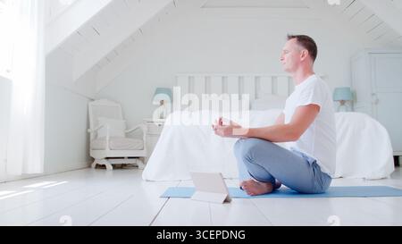 Mature Man With Digital Tablet Sitting In Bedroom Doing Yoga Stock Photo