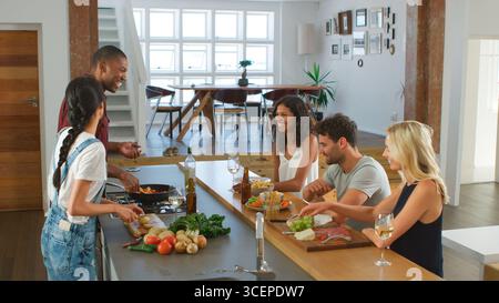 Couple Talking To Guests Whilst Making Food For Dinner Party At Home In Modern Kitchen Stock Photo