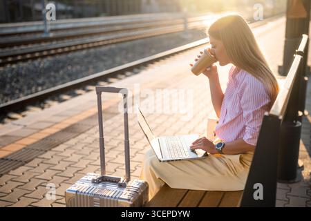 Elegant businesswoman using laptop and drinking coffee while sitting on a bench with suitcase at the railway station and waiting arrival of train Stock Photo