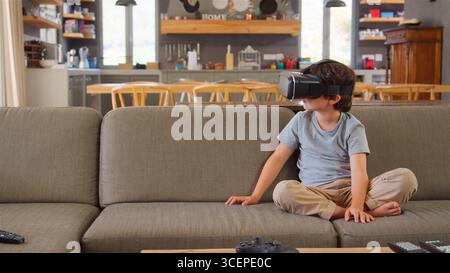Boy Sitting On Sofa At Home Wearing Virtual Reality Headset Stock Photo