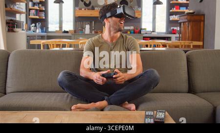 Man Sitting On Sofa At Home Playing Computer Game Using Virtual Reality Headset Stock Photo
