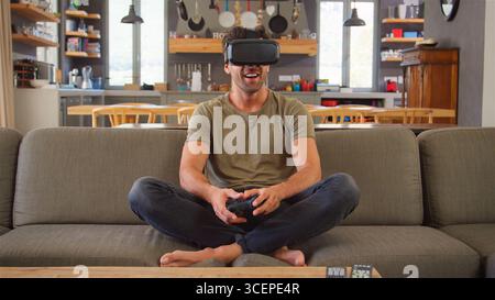 Man Sitting On Sofa At Home Playing Computer Game Using Virtual Reality Headset Stock Photo