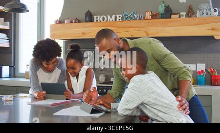 Parents Helping Children With Homework Using Digital Tablets In Kitchen Stock Photo