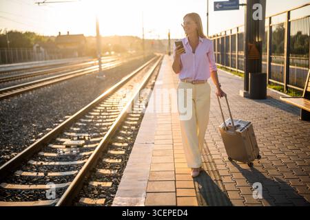 Elegant businesswoman walking confidently with suitcase and using smartphone at the sunny railway station. Stock Photo