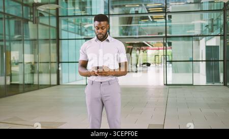 Businessman Standing In Lobby Of Busy Modern Office Using Mobile Phone Stock Photo
