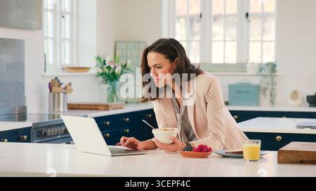 Businesswoman At Home In Kitchen Working On Laptop Whilst Eating Breakfast Stock Photo