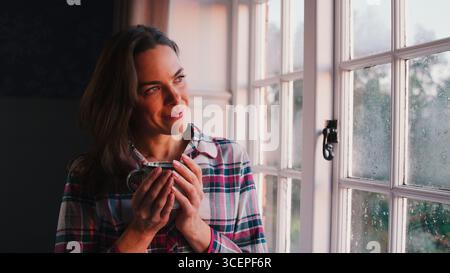 Woman At Home Looking Out Of Window Holding Hot Drink Stock Photo