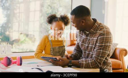 Father At Home Helping Daughter In Lounge As She Does Homework Using Digital Tablet Stock Photo