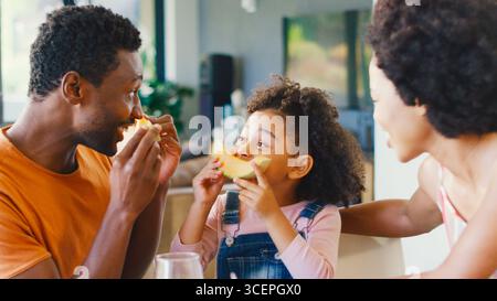 Family Shot With Parents And Daughter At Home Playing Game Making Face With Melon At Breakfast Stock Photo