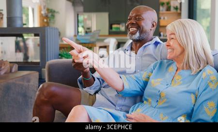 Multi-Racial Senior Couple On Sofa At Home Together Watching TV Stock Photo