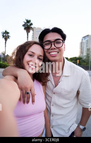 Multiracial couple using phone to take a selfie looking at camera smiling standing outdoors. Stock Photo
