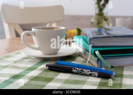 Cozy morning scene with white cup of coffee on saucer, stack of colorful notebooks, pens, and green checkered tablecloth on wooden table, blurred Stock Photo