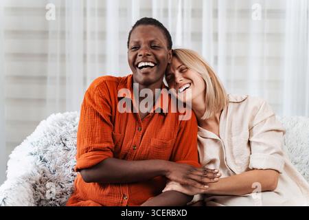 Two happy multi-ethnic friends laughing and embracing on the couch Stock Photo