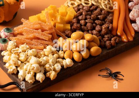 Halloween sweet charcuterie board with pumpkin, popcorn, dried fruits, color chocolate candy, spider, crackers on orange background. Festive food for Stock Photo