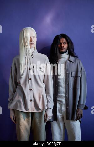 Vertical medium long studio shot of androgynous young Caucasian woman with albinism and handsome long-haired Indian man wearing casual clothes posing on camera, indigo blue background Stock Photo