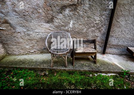 Two rustic chairs—one wicker and one wooden—sit side by side against an old textured wall on a mossy concrete surface. A quiet, nostalgic outdoor scen Stock Photo