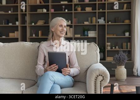 Smiling older lady relax on couch look aside from tablet Stock Photo