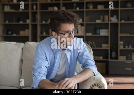 Unhappy young man sit on couch at home in silence Stock Photo