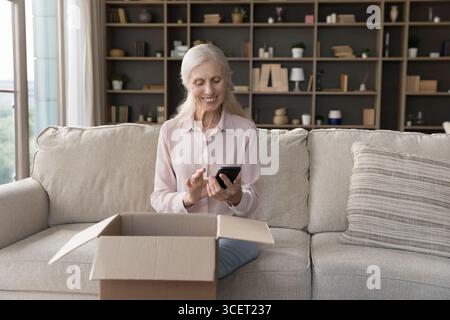 Elderly lady using smartphone sitting on sofa by opened delivery Stock Photo