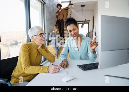 Mentorship and diversity in a modern office as colleagues share a laptop for team collaboration Stock Photo