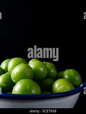 Close-up view of juicy green plums in a white bowl with blue rim, isolated on a black background. Stock Photo