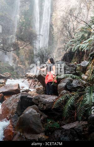 Indonesian woman seen from behind raising arms in freedom while standing near a tropical waterfall. Stock Photo
