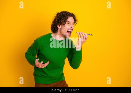 Curly-haired young man shouting into a mobile phone with an expressive gesture against a vivid yellow background. Stock Photo
